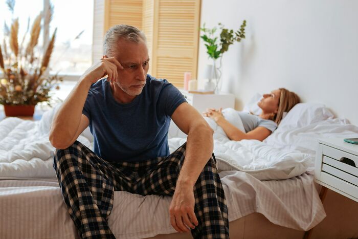 An older man sits on the edge of a bed looking distressed, with his wife sleeping in the background, representing truths about being married.