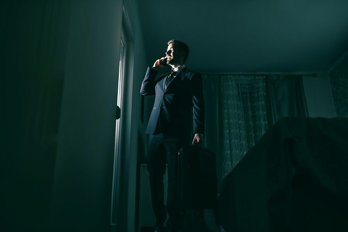 A man in a suit talks on a phone in a dimly lit room, luggage in hand, suggestive of scary true stories and questioning reality.