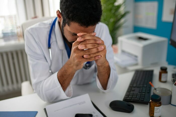 A doctor with a stethoscope sits at a desk with his head in his hands, reflecting the difficulty of delivering diagnoses.