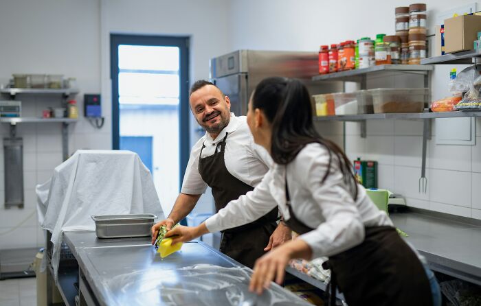 Two smiling coworkers cleaning a commercial kitchen counter, emphasizing personal details and workplace relationships.