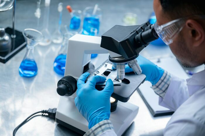 A scientist in gloves and safety goggles looking into a microscope in a laboratory with blue liquids. Scary science facts.