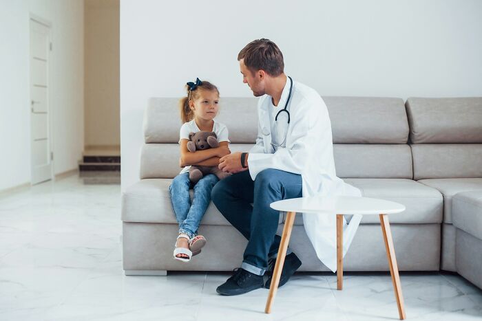 A male doctor speaking with a young girl holding a teddy bear, discussing a hard diagnosis.