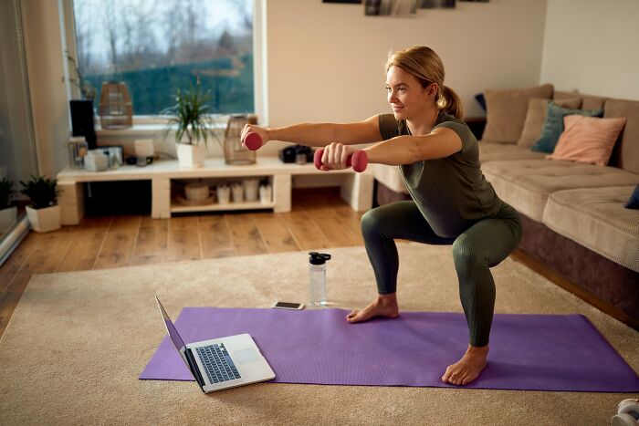 A woman doing squats with dumbbells on a yoga mat, with a laptop open nearby, showing a home workout.