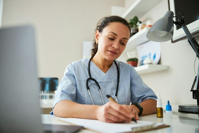 A female medical field worker in scrubs and a stethoscope writes on a clipboard at a desk with a laptop.