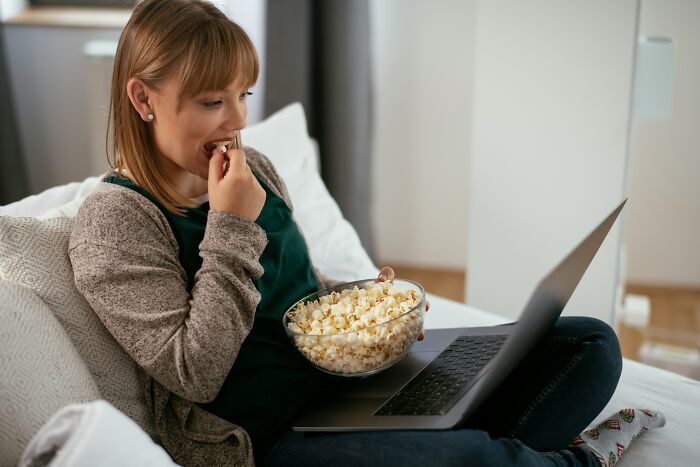 A woman eats popcorn while watching something on her laptop in bed, revealing a secret.
