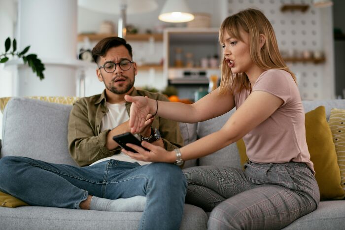 A couple arguing on a couch, the woman pointing at a phone held by the man, representing dumb reasons couples divorce.