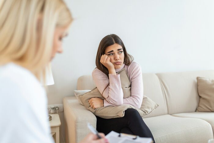 A sad young woman holds a pillow while sitting on a couch, looking away from her therapist during a therapy session.