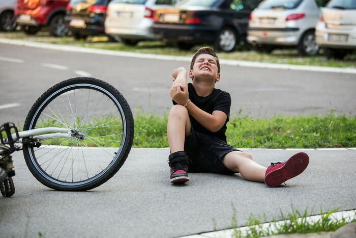 A boy sits on the ground next to his fallen bicycle, clutching his arm in pain, a horrible parent's neglect.