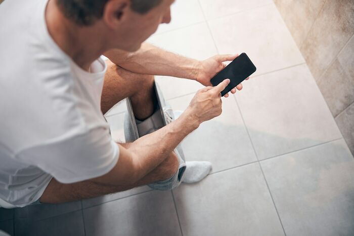A man sitting on a toilet, looking at his phone. An example of interesting neighbors and their habits.