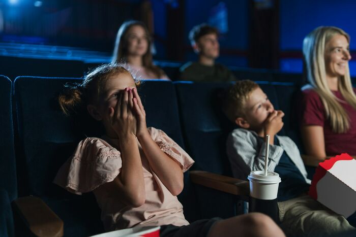 A young girl covers her eyes in a movie theater while a boy watches, reflecting horrible parent choices.