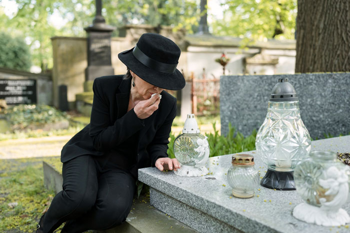 A woman in a black hat and dress wiping away a tear while kneeling at a gravestone in a cemetery, delivering a final wish at a funeral. A woman in a black hat and dress wiping away a tear while kneeling at a gravestone in a cemetery, delivering a final wish at a funeral.