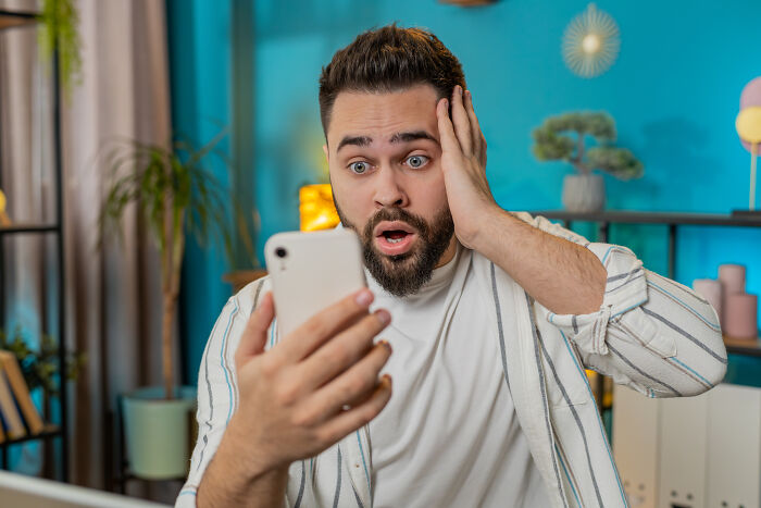 A surprised man with a beard holding a smartphone, looking shocked by what he sees, experiencing a sign from the universe.