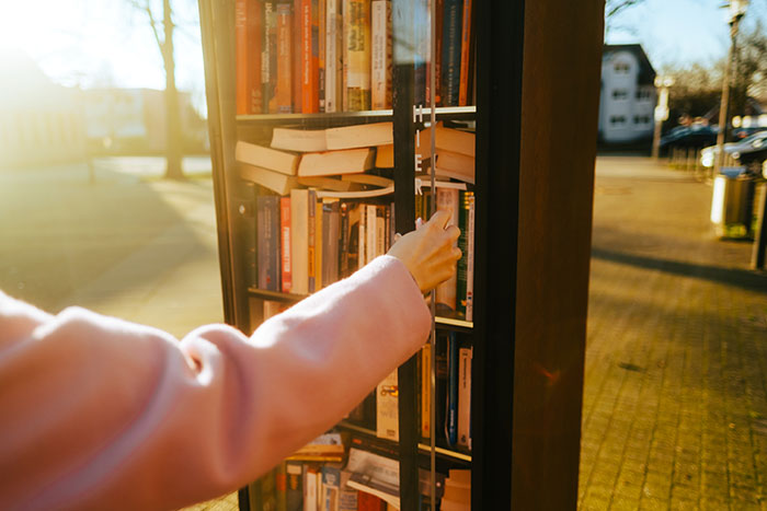 A hand opening a public bookcase filled with books under warm sunlight, sharing harmless secrets that warm your heart.