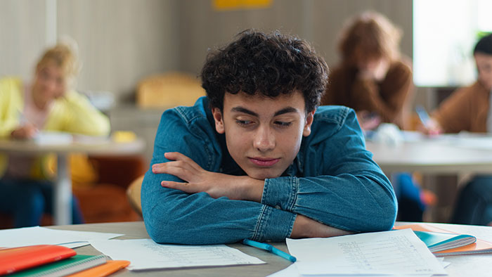 A young boy with curly hair resting his head on his arms at a desk, contemplating harmless secrets.