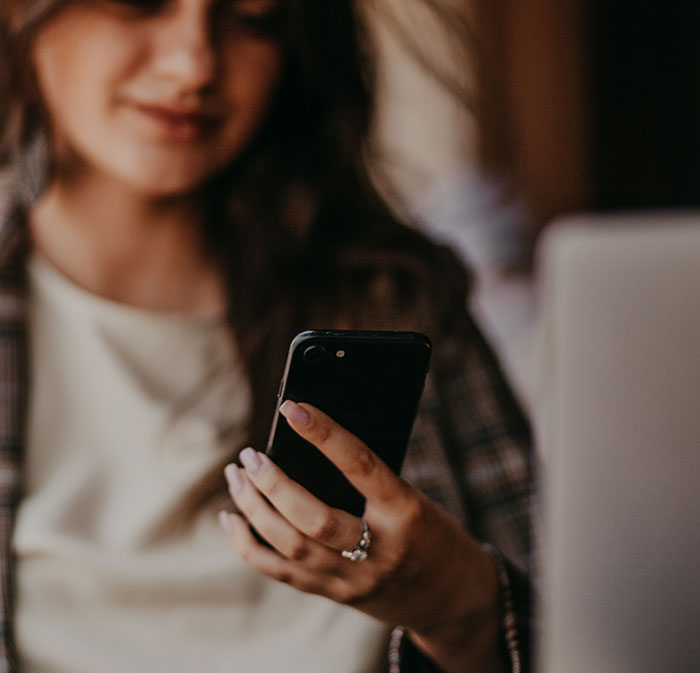 A woman holding a smartphone, with a laptop in the background, sharing a harmless secret that warms hearts.