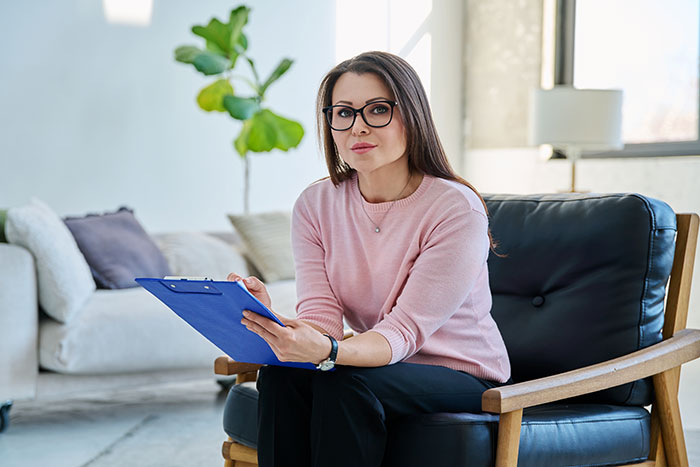 A woman in glasses and pink sweater sitting in a chair holding a clipboard, ready to hear harmless secrets.