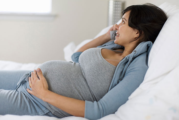 A pregnant woman resting in bed, cradling her baby bump while talking on the phone, a moment of secret joy.