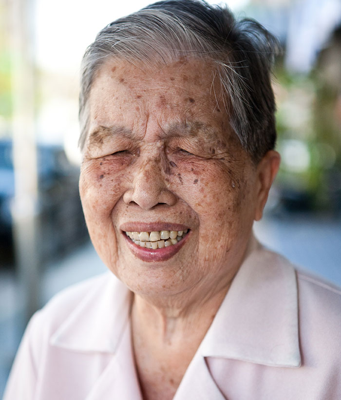 An elderly woman with a wide, warm smile and closed eyes, sharing a heartwarming secret.