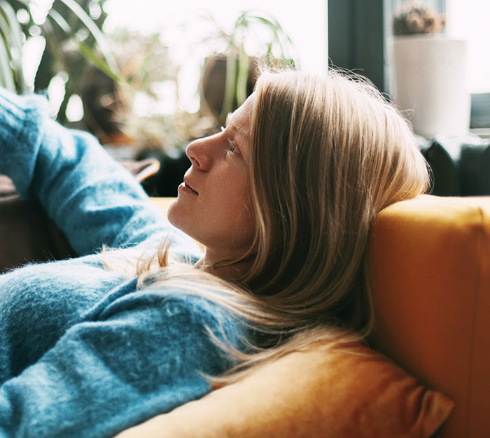A blonde woman relaxing on a yellow couch, gazing upwards thoughtfully, contemplating harmless secrets that warm your heart.