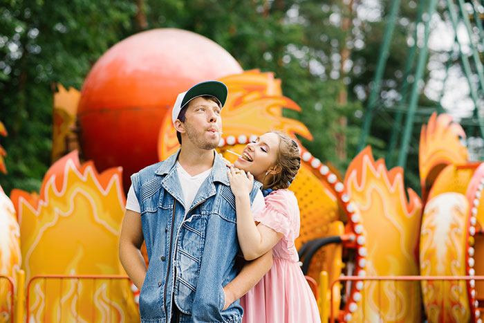 A couple at an amusement park with the woman playfully licking a lollipop held by the man, sharing harmless secrets.