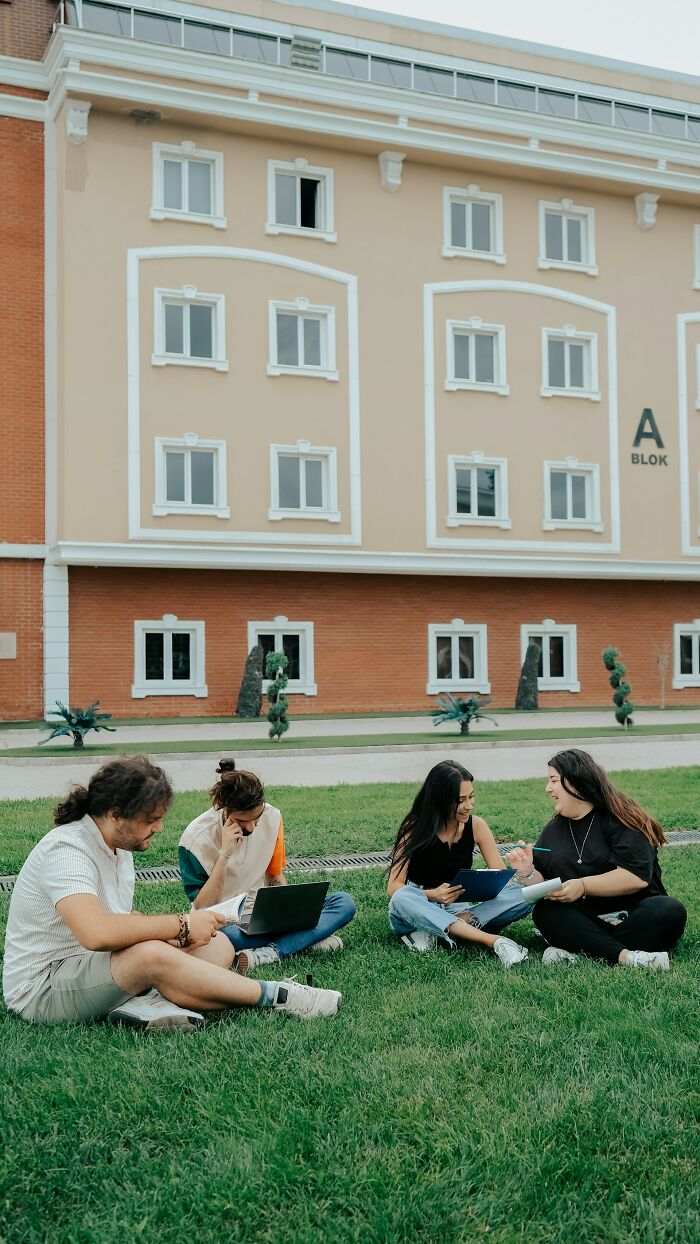 Four college students sit on a grassy lawn in front of a campus building, enjoying their new world of college life.