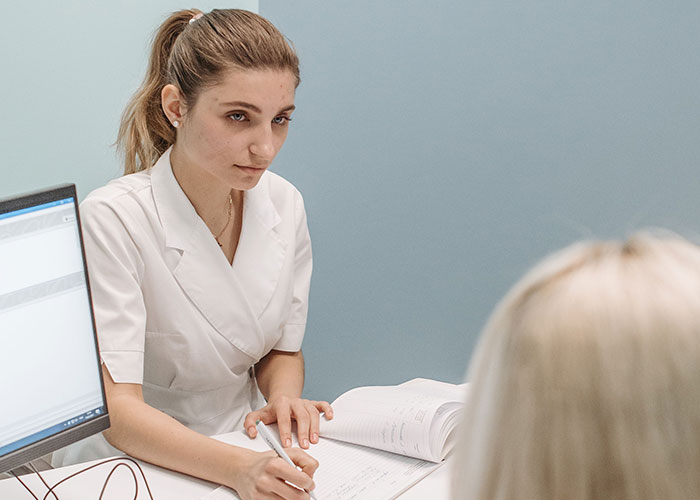 A female doctor in a white lab coat is attentively taking notes, listening to a patient about their symptoms.