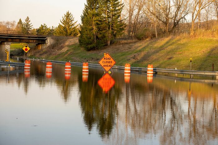 A flooded road with a 'Road Closed Ahead' sign, orange cones, and reflections. Harmless-looking things can be more dangerous.