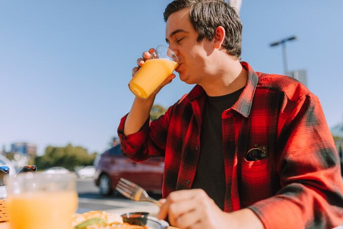 A man in a red plaid shirt sips orange juice at an outdoor table, recalling funny encounters with American tourists.