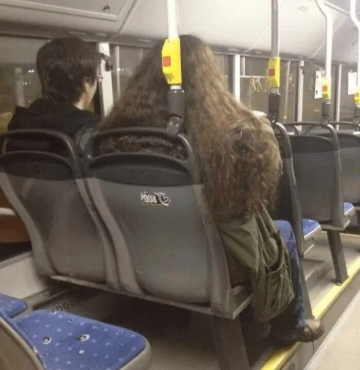 A person with extremely long, curly hair occupying two seats on a bus, a visual example of British humor.