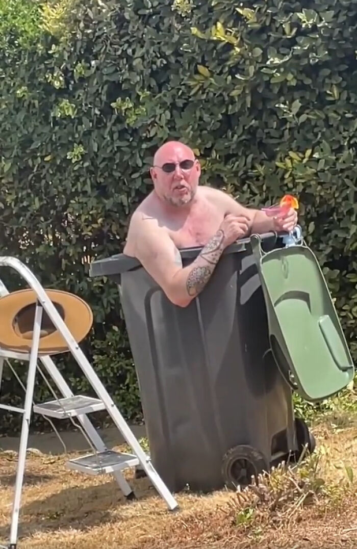 A bald man in sunglasses sits in a large black bin outside, exhibiting dry British humor.