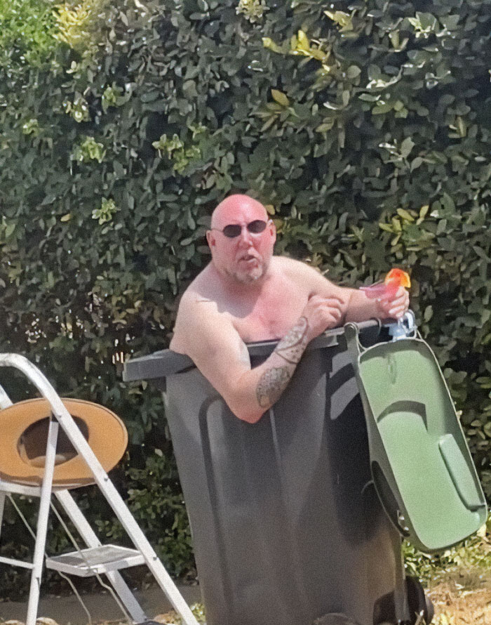 A bald man in sunglasses sits in a large black bin outside, exhibiting dry British humor.