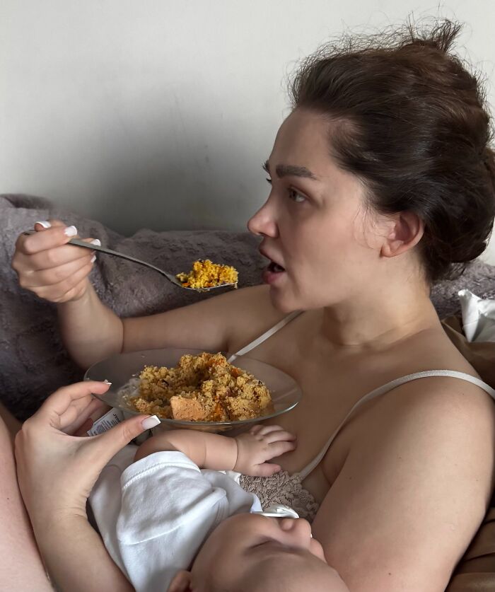 A mom eating while holding her baby, illustrating the joyful and fulfilling, but not easy, life of motherhood.