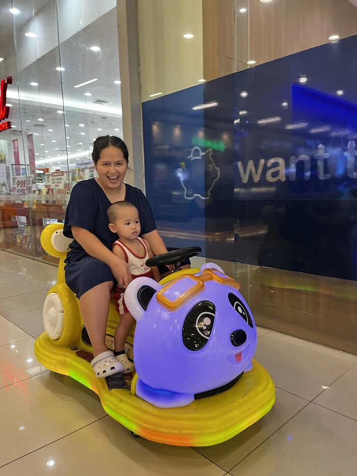 A smiling mom sits with her baby on a yellow and purple panda-shaped ride, illustrating the joyful side of motherhood.