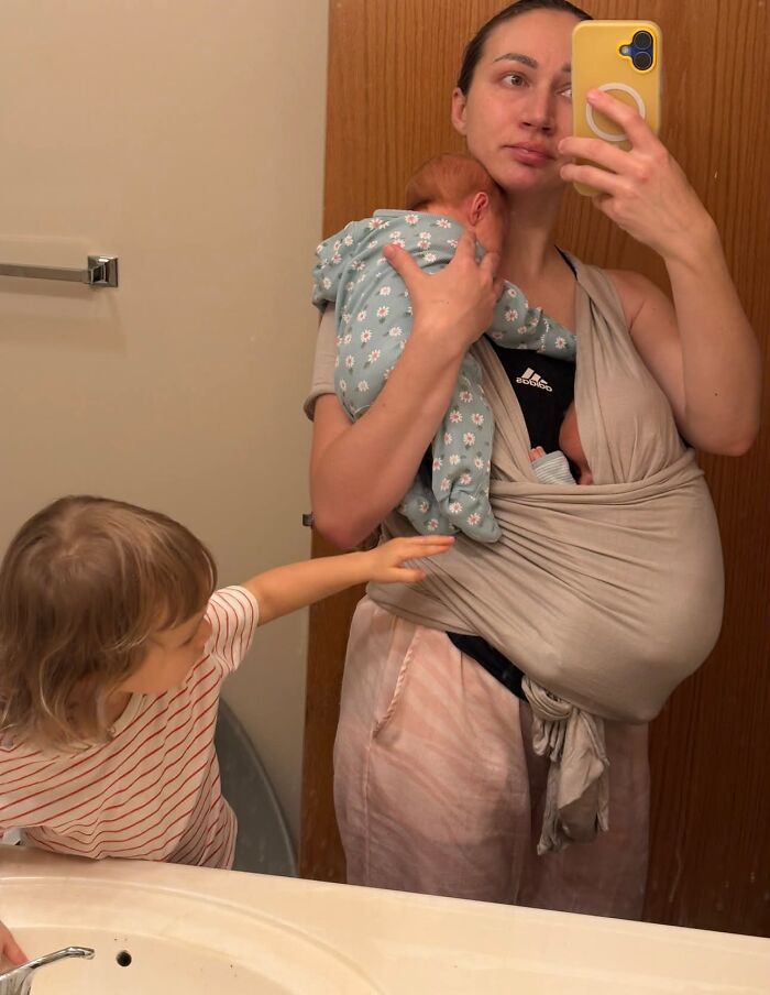 A mom holding a baby in a carrier, with another child reaching out, reflecting the demanding yet rewarding nature of motherhood.