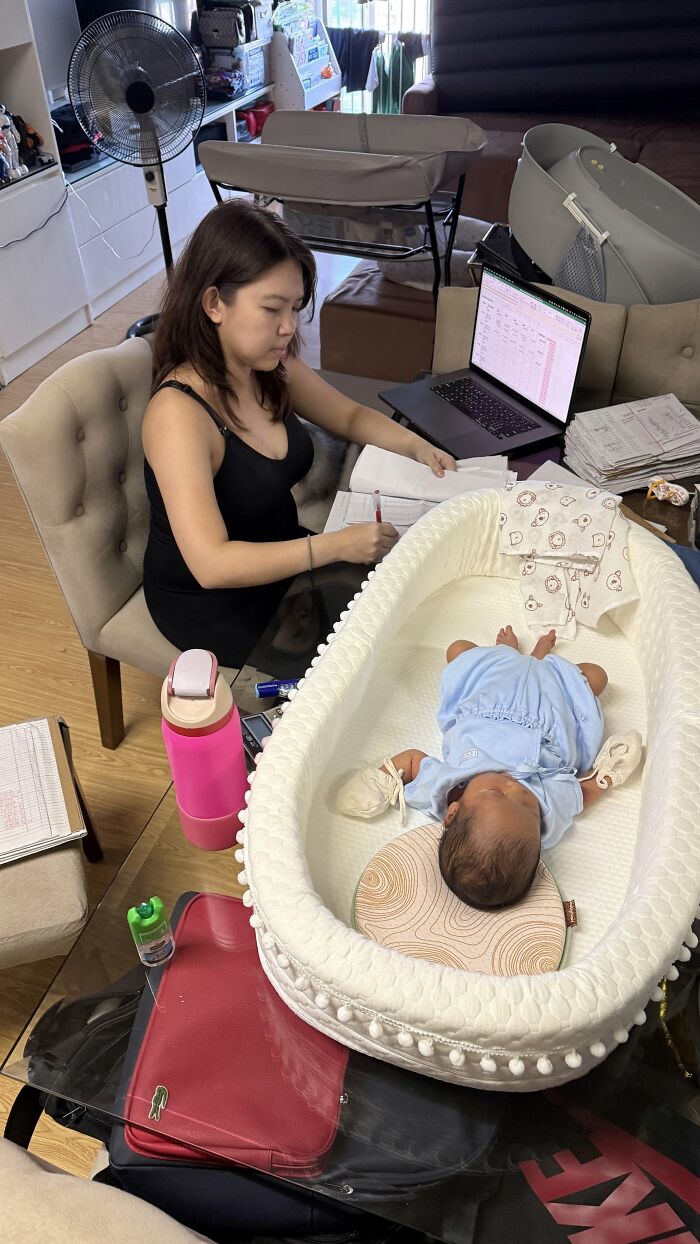 A mom works at a laptop while a baby sleeps in a bassinet nearby, illustrating the rewarding but not easy life of motherhood.