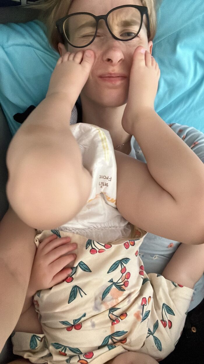 A mom with a baby on top of her, hands covering her face, capturing the joyful and sometimes messy reality of motherhood.