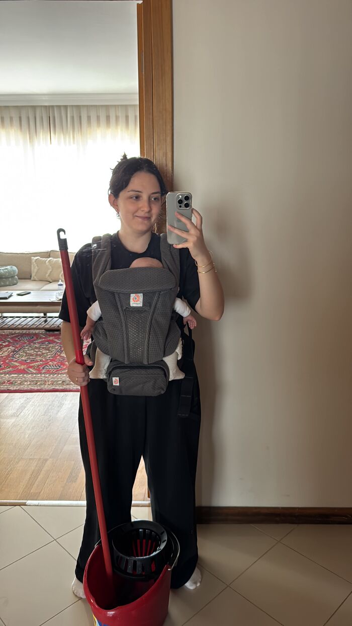 A mom with a baby in a carrier and a mop, illustrating that motherhood is rewarding but not easy.