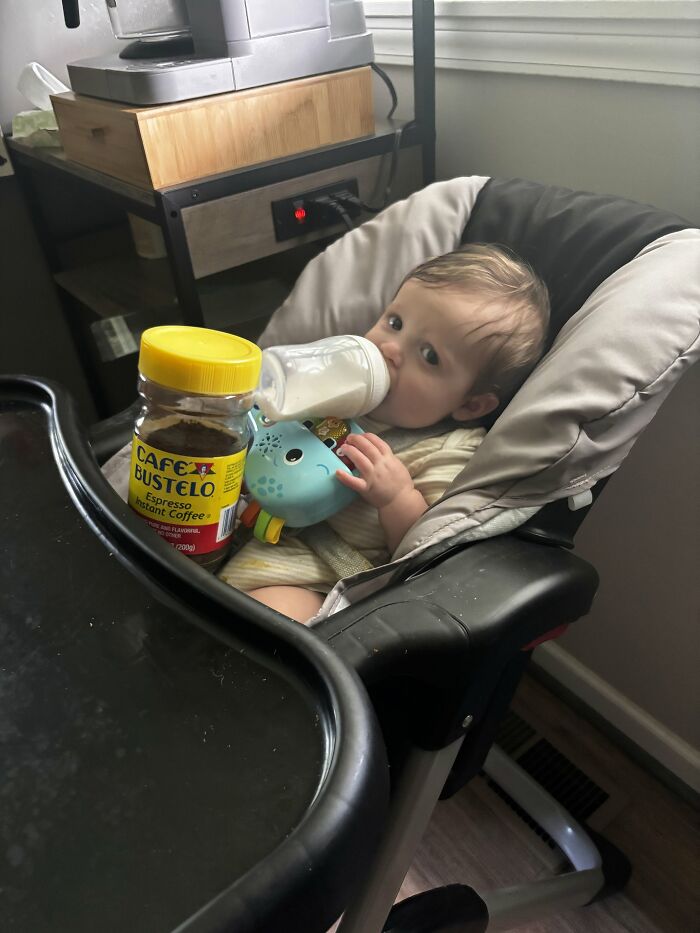 A baby in a high chair drinking from a bottle, next to a jar of coffee, illustrating the demands of motherhood.