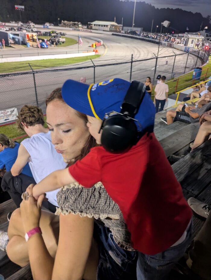 A mom hugs her child wearing a hat and ear protection at a racetrack, showing a rewarding motherhood moment.