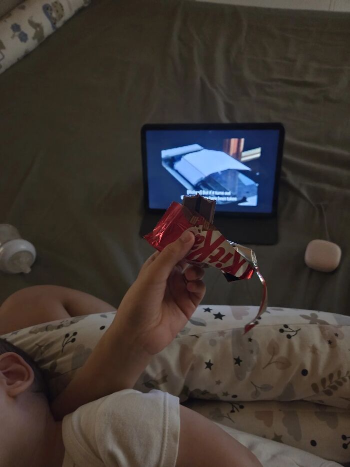 A mom eats a Kit Kat bar while lying down next to a sleeping child, representing the not easy part of motherhood.
