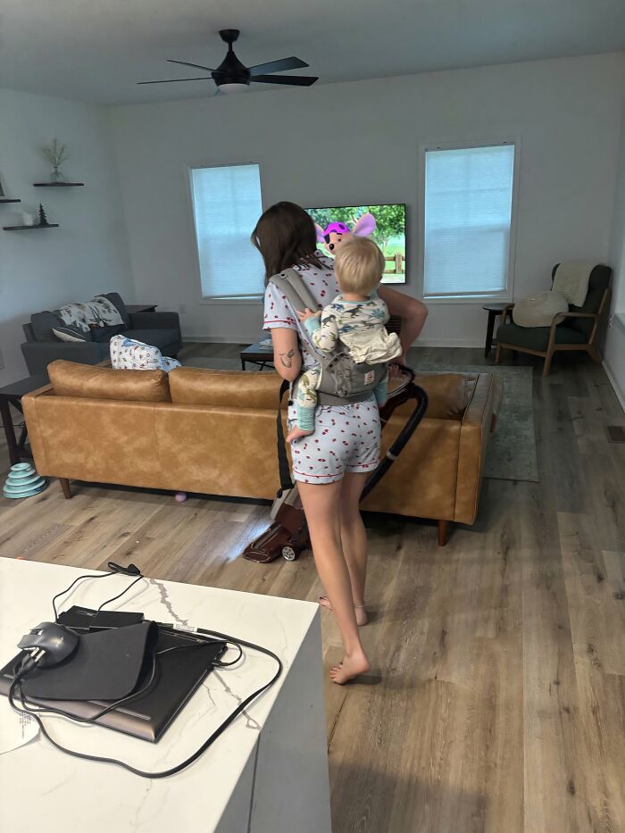 A mom vacuums with a baby in a carrier on her back, illustrating the fulfilling but not easy side of motherhood.