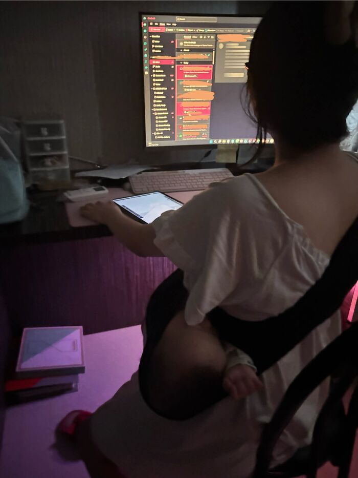 A mom working at a computer with her baby sleeping in a carrier on her lap; motherhood can be challenging but rewarding.