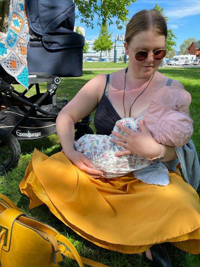 A mom in sunglasses and a yellow skirt is breastfeeding her baby on a sunny day in the park; motherhood is fulfilling.