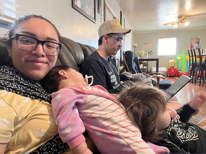 A mom with glasses smiling at the camera, holding a sleeping baby while another child sits beside them; motherhood is rewarding.