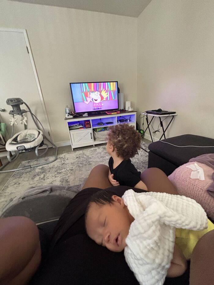 A baby sleeps on a mom's lap while an older child watches TV, highlighting the joyful aspects of motherhood.