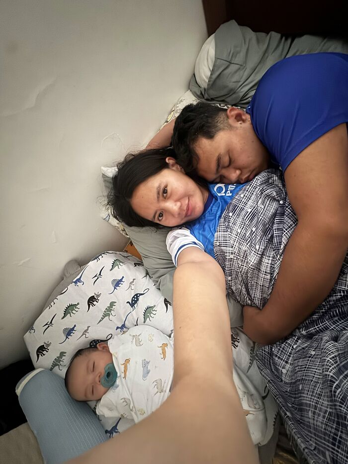 A mom smiling at the camera while her partner and baby sleep beside her in bed, depicting rewarding and joyful motherhood.
