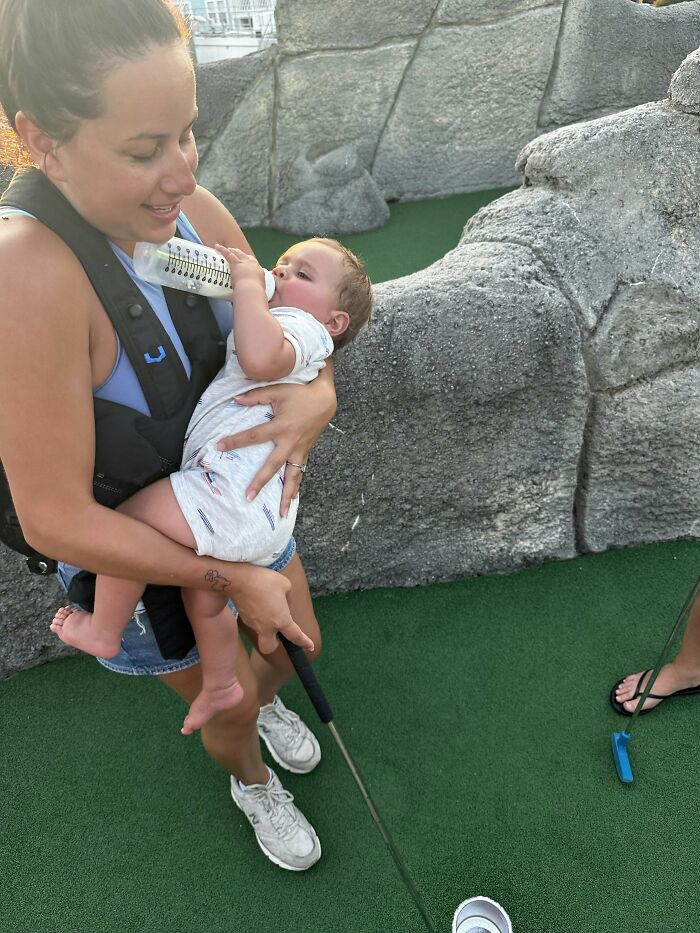 A mom feeds her baby a bottle while playing miniature golf, showing a rewarding motherhood moment.