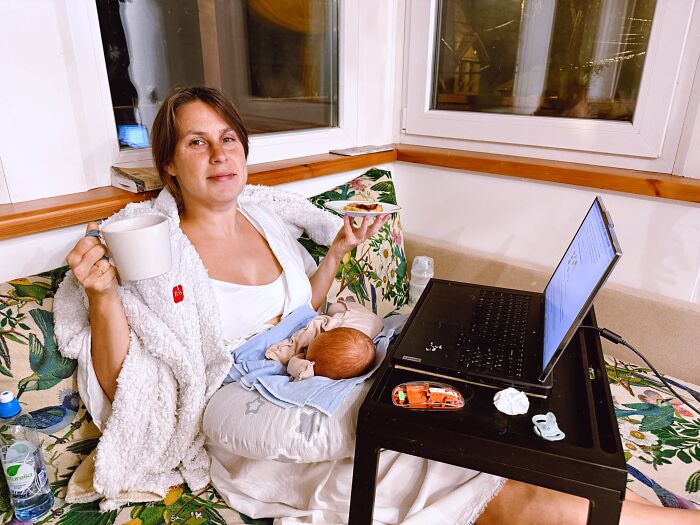 A mom relaxing on a couch, holding a mug and a snack, with her baby beside her and a laptop on a tray, depicting rewarding motherhood.