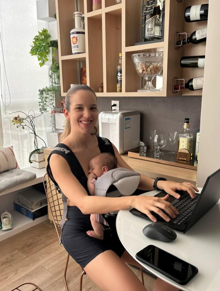 A smiling mom working on a laptop with her baby in a carrier, showcasing the rewarding and fulfilling aspects of motherhood.