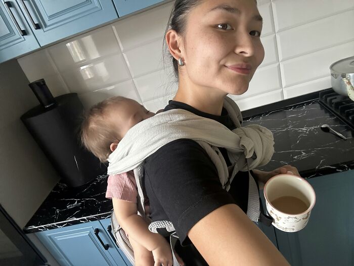 Motherhood is rewarding: A mom multitasking, carrying her baby in a wrap while holding a cup, showing a fulfilling moment.
